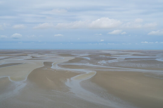 Aerial view of tidal flats with winding water channels and wet sand. Sky has scattered white clouds over calm horizon. Scene shows natural coastal landscape without human presence