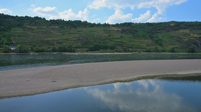 Ein Fluss mit einer Sandbank und &uuml;ppigen H&uuml;geln, die teilweise mit Weinbergen bedeckt sind, unter einem klaren, blauen Himmel, Bacharach am Rhein, Rhein, Rheingau, Hessen, Deutschland