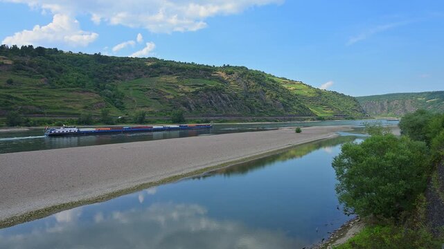 Ein Fluss mit einer Sandbank und &uuml;ppigen H&uuml;geln, die teilweise mit Weinbergen bedeckt sind, unter einem klaren, blauen Himmel, Bacharach am Rhein, Rhein, Rheingau, Hessen, Deutschland