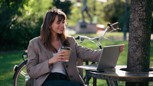 Video of beautiful woman working on a laptop while drinking coffee in the park.