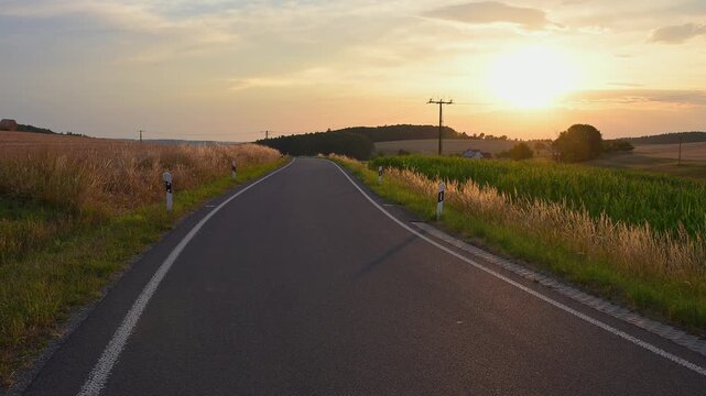 Eine Landstra&szlig;e, die sich durch die Felder schl&auml;ngelt, w&auml;hrend die Sonne am Horizont untergeht, Sommer, Reichartshausen, Amorbach, Odenwald, Bayern, Deutschland