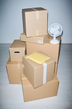 Vertical shot of cardboard boxes and padded mailer stacking indoors beside small fan, suggesting packing, storage, relocation, shipping workflow. Ideal for e commerce, delivery, logistics, moving ads