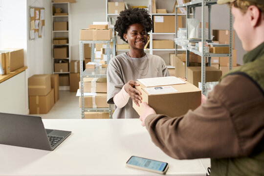 Young Black woman handing parcel to customer at fulfillment counter, processing order pickup in small warehouse. Ideal for e commerce, shipping service, retail logistics marketing
