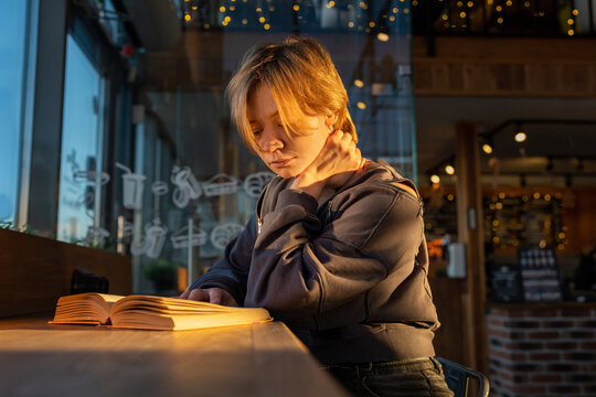 Young reader by window in cozy cafe interior, warm evening light, reflective glass, wooden counter, soft string lights, and quiet focus creating calm urban atmosphere