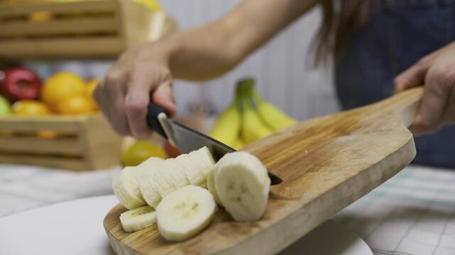 Cinematic slow motion of woman using knife to scrape banana slices off cutting board and onto plate, closeup