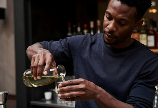 African american bartender pouring liquid from a glass carafe into a jigger at a bar counter, the bar counter setting includes blurred bottles in the background, indicating a professional hospitality