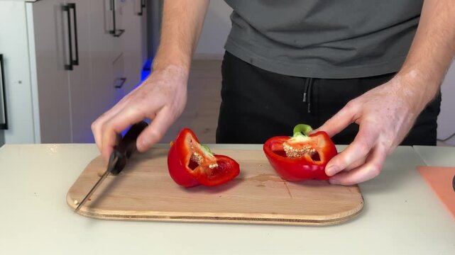Close view of hands chopping red bell pepper on wooden cutting board in bright modern kitchen, knife slicing through fresh vegetable with seeds, stem, and clean preparation detail