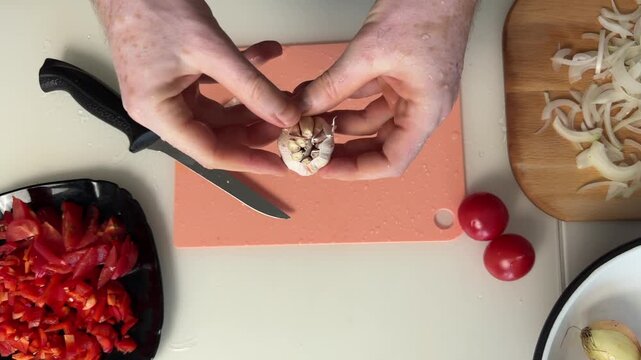 Overhead hands peeling garlic cloves on peach board beside chopped peppers onions and tomatoes, with chef knife and bowls in bright home kitchen prep scene, with garlic, hand, peeling, clove detail