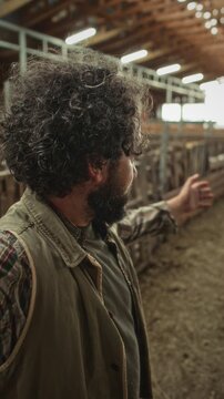 Middle-aged male farmer with curly hair and beard waving hello and speaking on smartphone camera while recording vlog or video calling inside livestock barn. Vertical POV shot