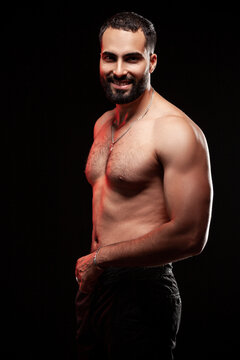 Smiling fit guy posing shirtless showing athletic biceps and strong abdominal muscles with warm red side lighting highlighting skin texture isolated over black background