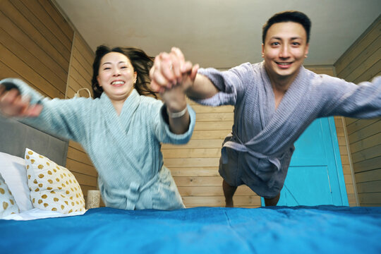 Joyful Couple Play on Bed Holding Hands in Robes, Happy Hotel Vacation