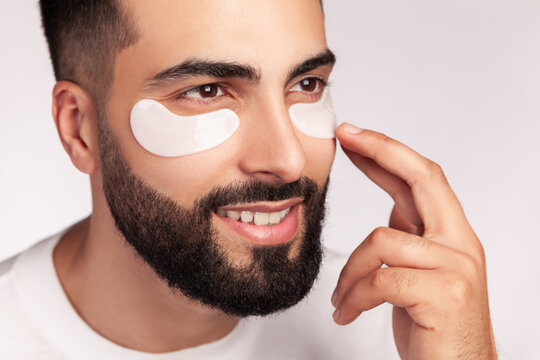 Guy wearing white hydrogel patches under eyes for beauty therapy and dark circles reduction procedure isolated over gray background