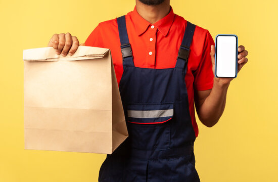 Delivery service mobile app. Deliveryman in overalls showing paper parcel and cellphone with blank display, mock up for express shipping advertise. Indoor studio shot isolated on yellow background.