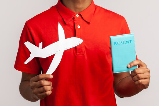 Satisfied man with happy expression wearing red casual style T-shirt, holding passport and paper plane in hands planning vacation abroad. Indoor studio shot isolated on gray background.