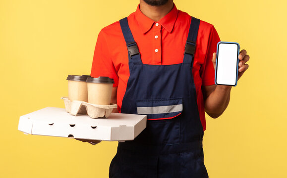 Delivery mobile app, internet advertise. Friendly courier man in overalls holding coffee cups and cellphone with mock up display, online order service. Indoor studio shot isolated on yellow background