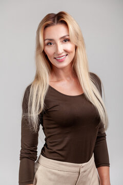 Cheerful blonde woman smiling widely at camera wearing brown casual long sleeve shirt and beige high waisted trousers standing isolated over gray background