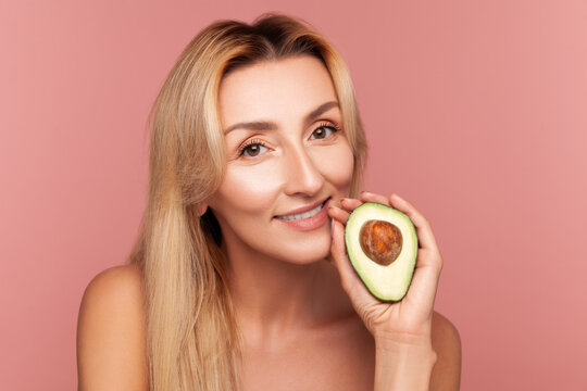 Smiling lady posing with half of ripe avocado near cheek, highlighting moisturizing benefits of natural oils for skin health isolated on pink background