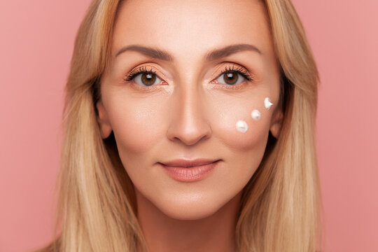 Close-up front view of blonde mature woman with three moisturizer dots applied under her eye looking at camera isolated on pink background