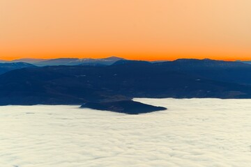 Naklejka premium A panoramic mountain view showing a sea of white clouds blanketing the valley below a vibrant orange horizon at twilight