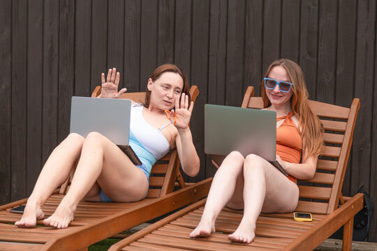 Two women using laptops on sun loungers in backyard