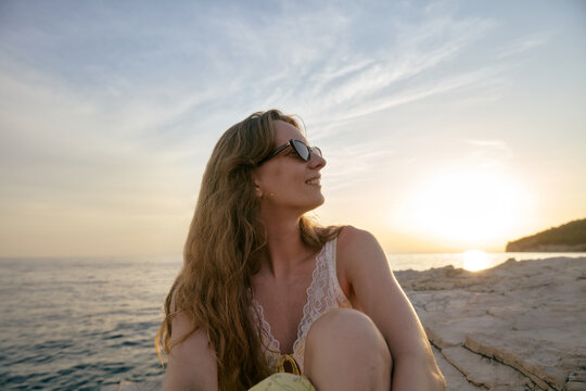 Young Woman Sitting on Rocky Shore Enjoying Sunset Over the Sea