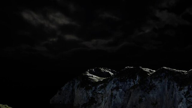 Cliffs rise steeply from the ocean, illuminated by some light. Dark clouds fill the night sky above creating a dramatic scene. The ocean is calm and mirrors the cliffs.