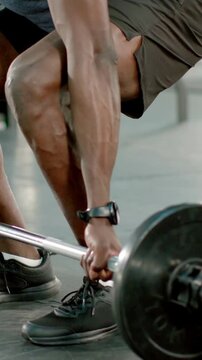 Vertical video: Gripping barbell, man in gray tee and black shorts deadlifting in gym, training