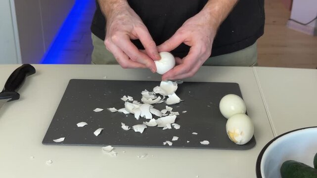 Hands peeling hard boiled egg over dark cutting board in home kitchen, close up scene with broken shell fragments, peeled eggs, cooking prep, and clean food arrangement