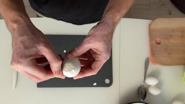 Overhead kitchen scene showing hands peeling hard-boiled egg over black cutting board on white countertop, with shells breaking away, soft natural light, and clean domestic preparation