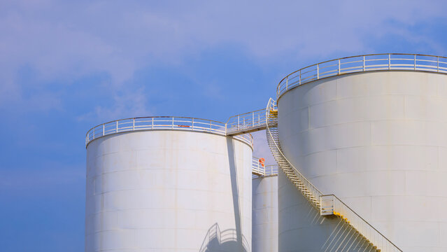 White industrial fuel storage tanks with metal stairs against blue sky background in oil refinery plant.