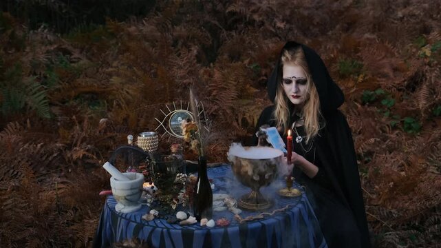 Seated woman in black hooded cloak and pale dramatic makeup holds card over steaming metal bowl while steadying lit candle nearby; vapor rises softly among autumn ferns and ritual objects