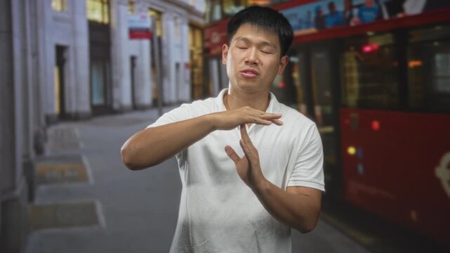 Man making timeout hand gesture with hands near chest on an urban street beside a red doubledecker bus and stone building facades; concentration.