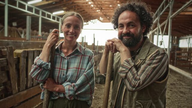Joyful male and female farmers leaning on their pitchfork handles, looking at camera and smiling during break from work inside wooden barn. Video portrait