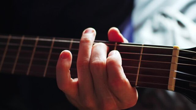 A person sits in a dimly lit room, focused on playing a guitar. The hand presses down on the strings