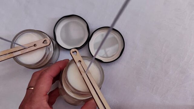 Woman trims a candle wick and removes a wooden wick holder from a soy candle on a white tablecloth, handmade finishing process, artisan production, DIY craft, product preparation.