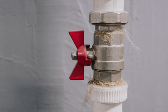 Metal ball valve with red butterfly handle installed on white plastic plumbing pipe indoors