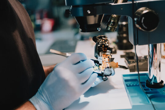 Camera repair technician servicing exposed electronic components of a disassembled digital camera under a stereo microscope at a technical workbench