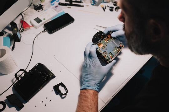 Camera repair technician inspecting exposed circuit boards inside a disassembled digital camera at an electronics service workbench