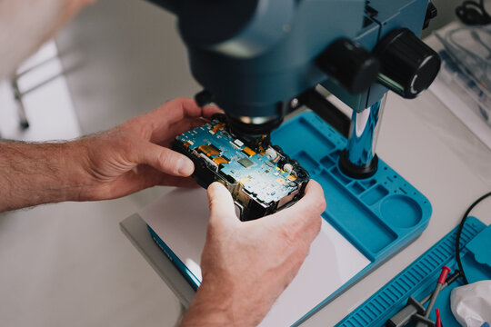 Electronics technician inspecting a disassembled digital camera circuit board under a stereo microscope at a repair workbench
