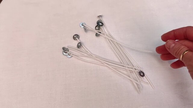 Woman selects a single candle wick from scattered supplies on a white tablecloth, candle making preparation, handmade craft, DIY workspace, minimalist lifestyle.