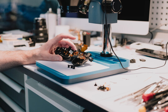 Camera repair technician positioning a disassembled digital camera under a stereo microscope for precision electronics inspection at a service workbench
