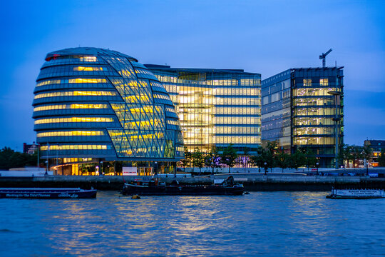 Night view of London's City Hall (the "onion" building) illuminated on the Thames, with an adjacent office tower and two moored boats marked "Port of London" and "Working for the Tidal Thames" in the 