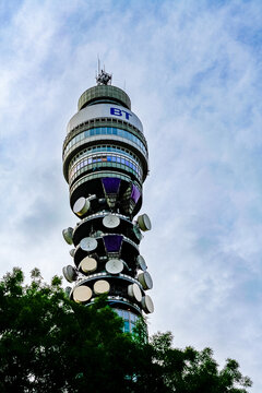 Low-angle view of the BT Tower in London, showing its rotating restaurant section, satellite dishes, and the "BT" logo, framed by tree branches against a cloudy sky.