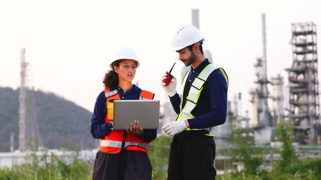 Two industrial engineers analyze data on laptop and radio communication at refinery, managing petroleum, chemical processes, and plant operations.