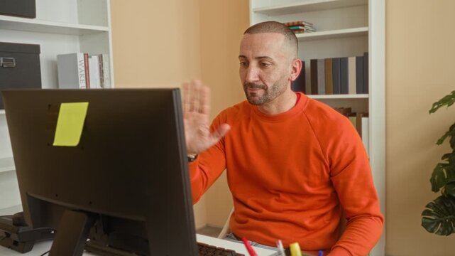 Man at computer waves hand to monitor while seated by bookshelves in building office with orange sweater visible; friendly interaction.