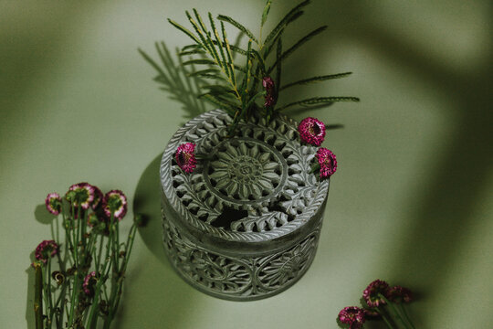 An ornate silver trinket box with a carved circular lid is surrounded by magenta flowers and green foliage, with dramatic botanical shadows crossing the muted green background.