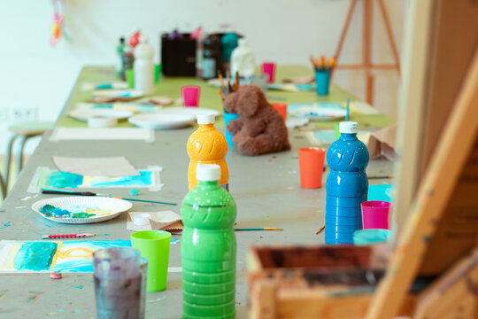 Colorful children's art workshop with paint bottles, brushes, and paper scattered across a table, featuring a teddy bear and various art supplies in a bright indoor setting