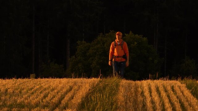 Cool male Hiker with backpack walking on a field at sunset. Warm orange tones standing out against dark background.