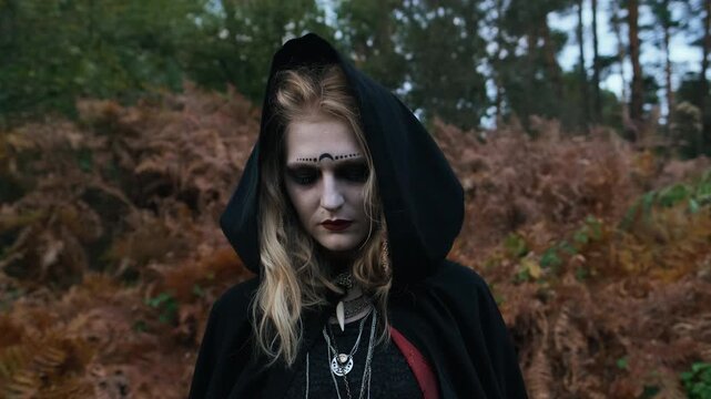 Head and shoulders view of young woman in black hooded cloak within outdoor woodland, surrounded by blurred autumn ferns, distant trees, pale makeup, face markings, and somber mystical mood