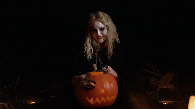 Centered medium close portrait of pale woman wearing dark clothing, heavy theatrical eye makeup, dark lipstick, and decorative forehead dots while gripping large glowing carved pumpkin in moody